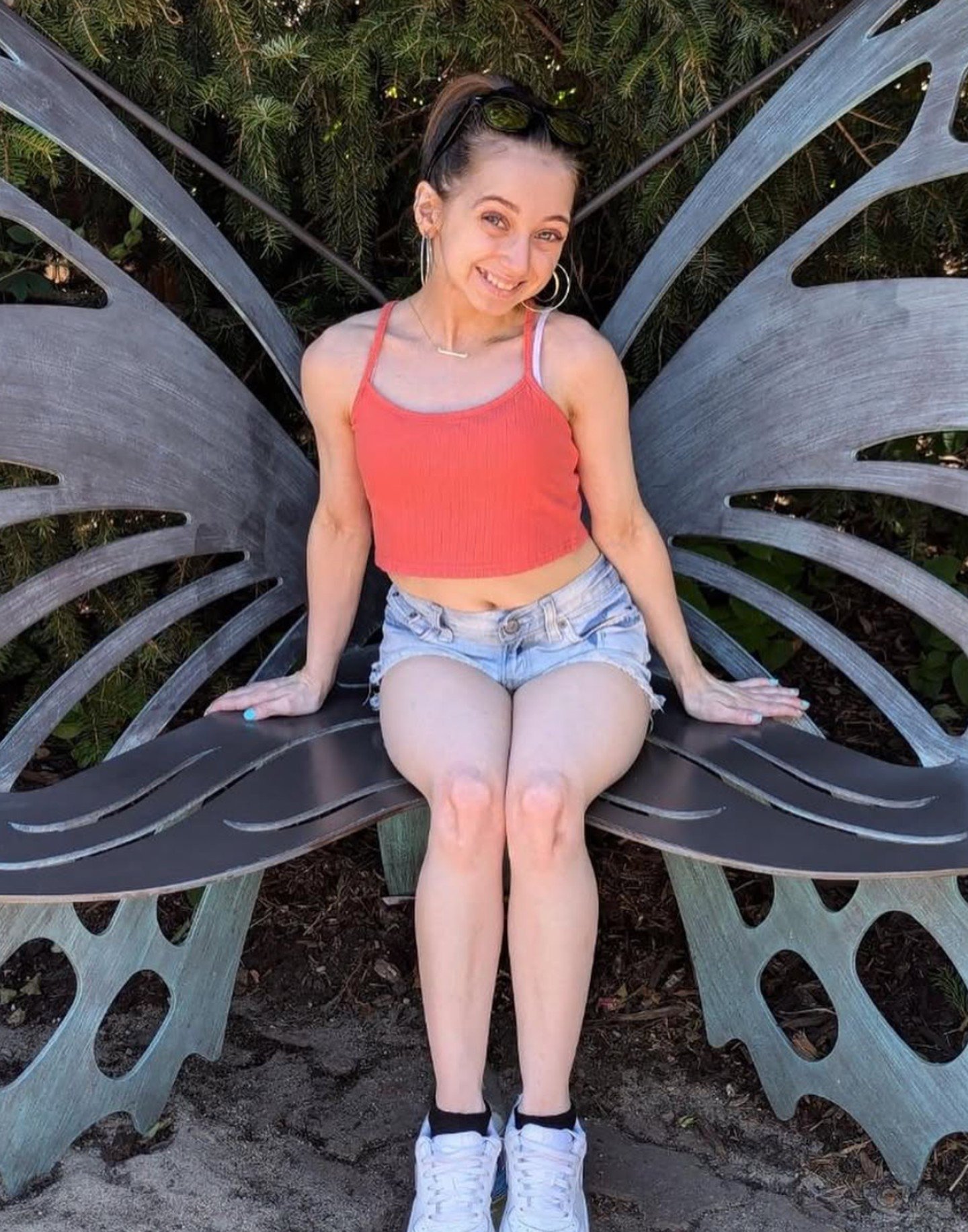 Shauna Rae in a red top and denim shorts sitting outdoors, smiling